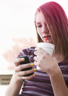 Bild på en flicka som dricker en läsk och använder sin mobiltelefon.